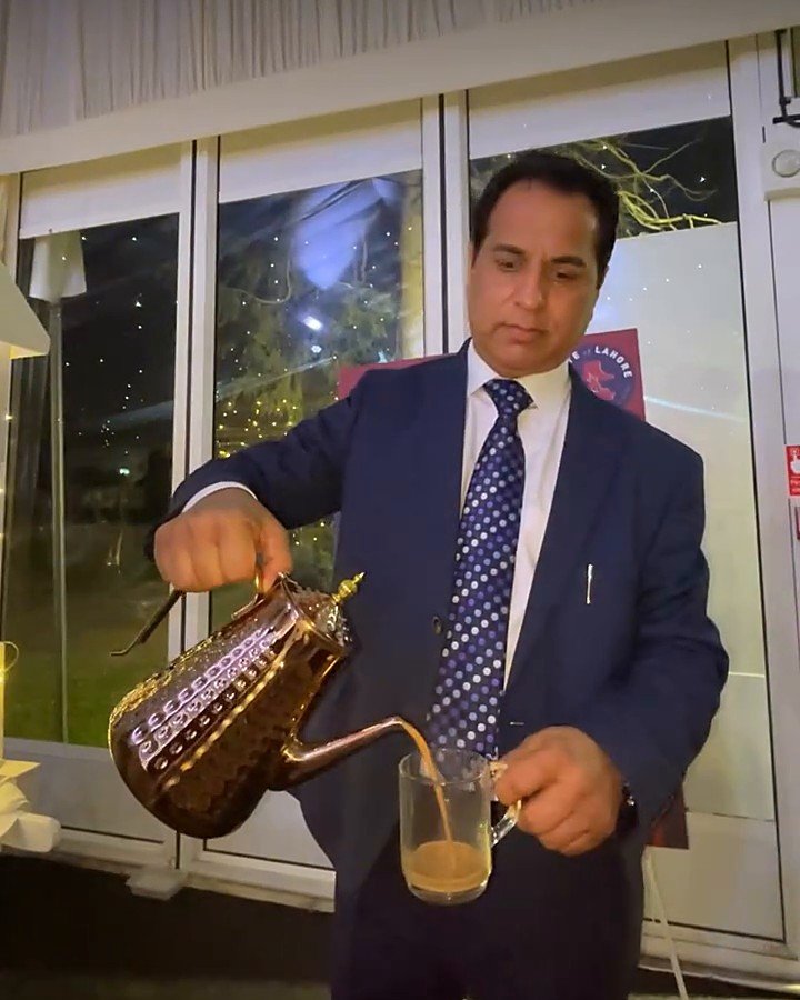 Taste of Lahore catering team member in a navy suit pouring karak chai from a brass pot